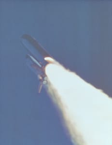 The Shuttle Challenger roughly 53 seconds into flight.  A fiery plume is visible on the right booster just above the rocket blast.  Credit: NASA.